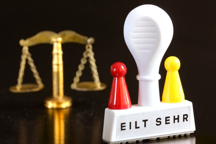 Die Aufnahme zeigt im Hintergrund die symbolisch Waage des Gerichts sowie vorne einen Stempel mit Aufschrift - Eilt sehr -